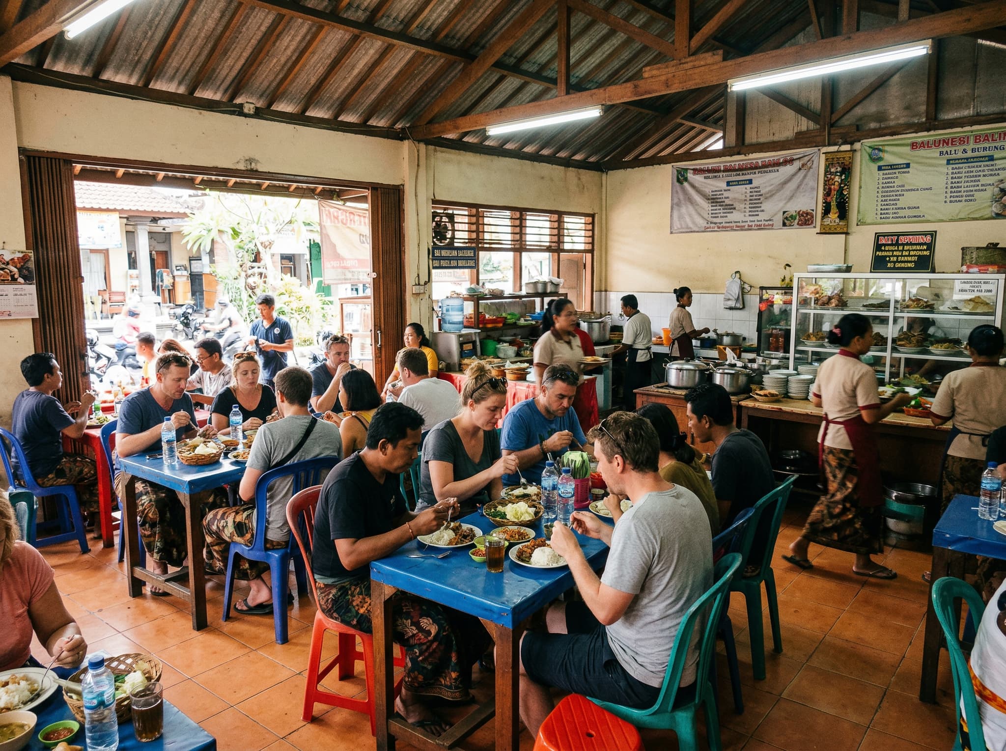 The interior of a traditional Balinese warung at midday — communal plastic tables and chairs, fluorescent lighting, diners eating babi guling from simple plates, capturing the no-frills setting described in the 'Practical Details' section