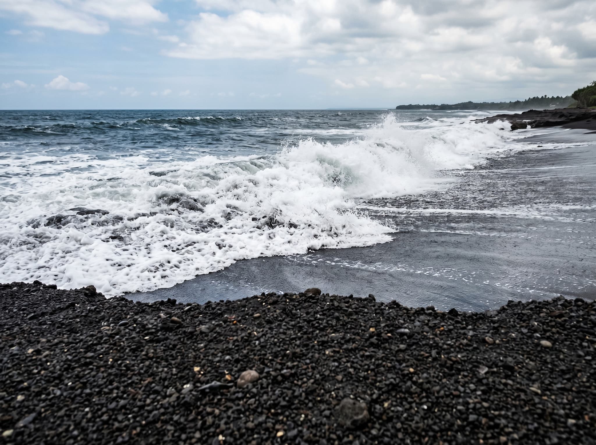 Ground-level view of Seseh Beach's black sand at midday, showing the coarse volcanic texture underfoot and strong surf breaking in the background — illustrating the beach's raw, unmanicured character described in the article