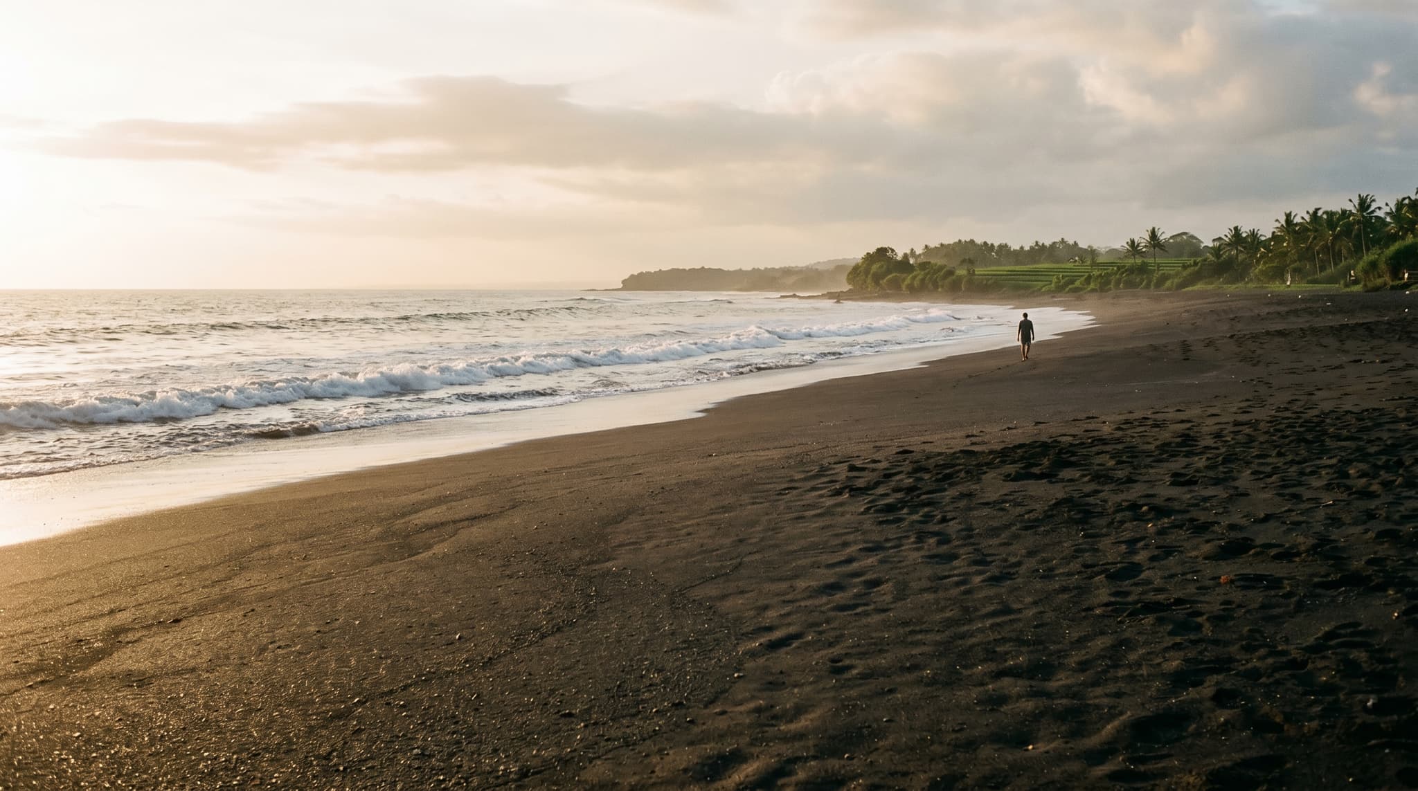 Seseh Beach's wide black volcanic sand shoreline at golden hour, with the dark surf breaking under a warm low sun — establishing the quiet, undeveloped character of this stretch of Bali's southwest coast