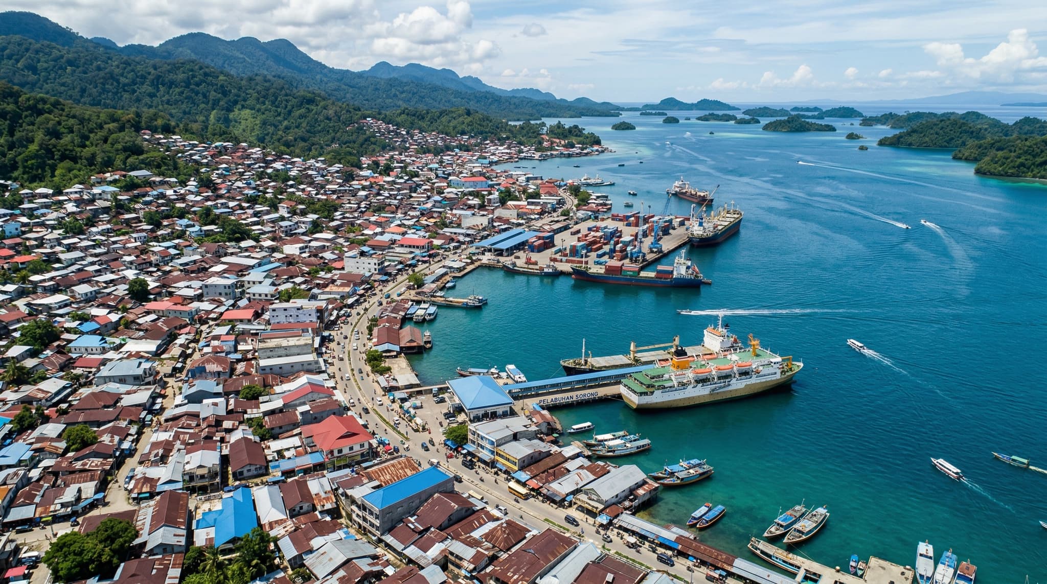 Aerial or waterfront view of Sorong city, West Papua, Indonesia — a dense coastal port city on the edge of New Guinea, establishing the article's premise of a functional transit hub with the sea visible, conveying the city's role as a gateway rather than a destination