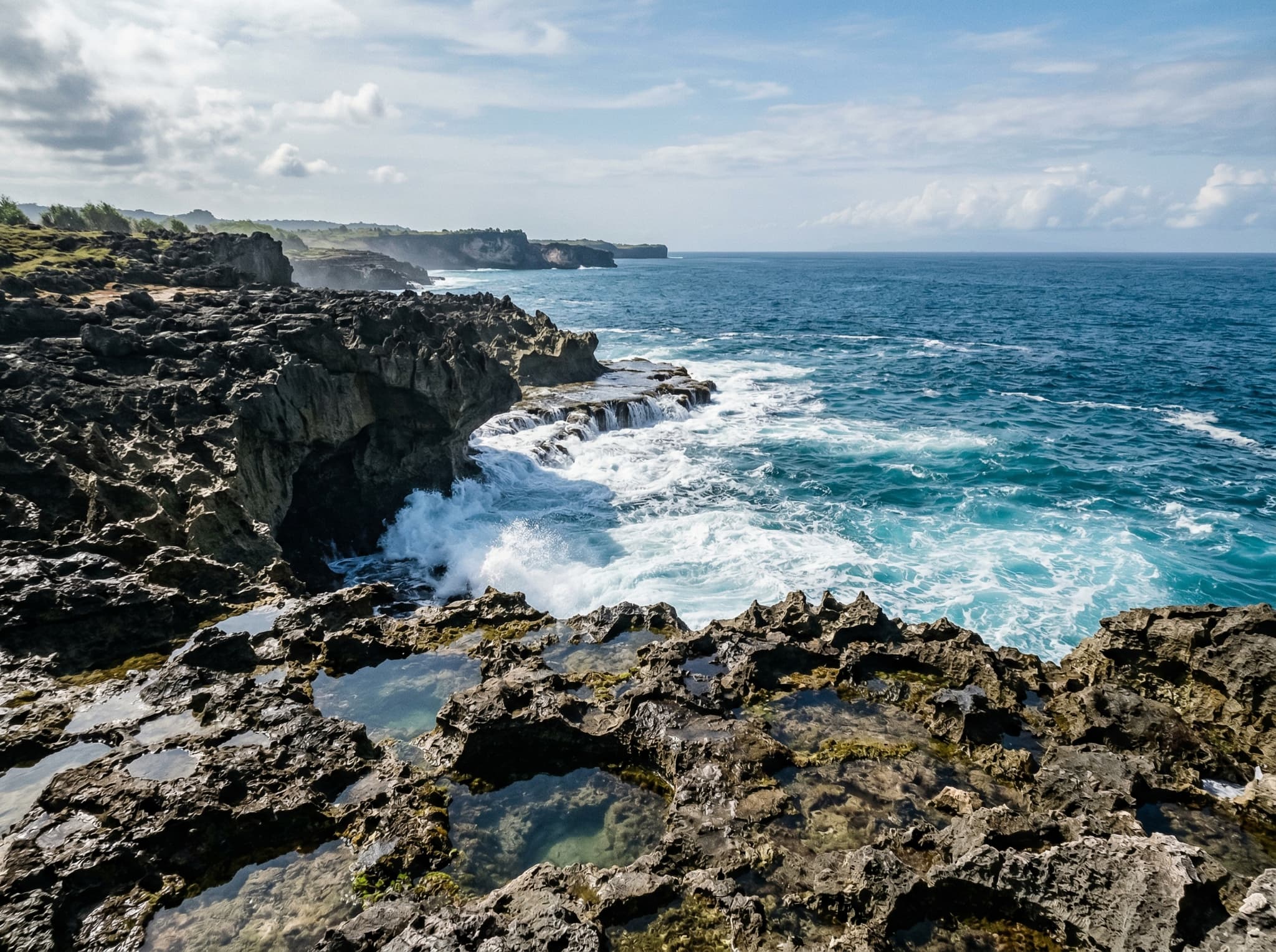 The rugged limestone coastline south of Devil's Tear on Nusa Lembongan — jagged rock shelves and tide pools extending along the cliff walk toward Sandy Bay, with waves breaking against undercut cliffs, showing the less-visited stretch the article recommends exploring beyond the main blowhole
