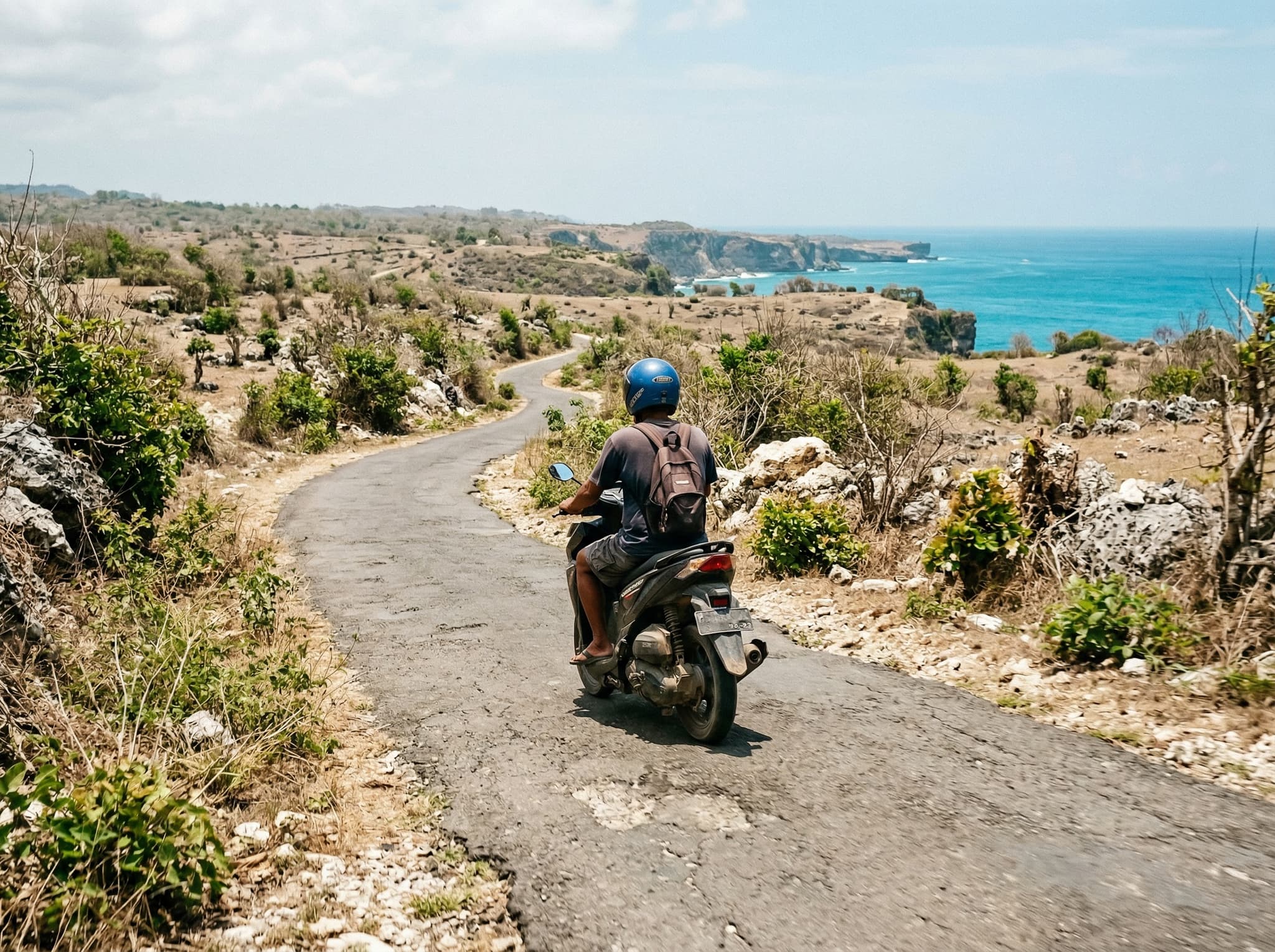 A scooter rider navigating a narrow cliff-top lane on the Bukit Peninsula with dry scrubby limestone vegetation on either side and a glimpse of ocean in the distance — illustrating the article's practical note that a scooter is the primary way to get around the peninsula's remote roads.