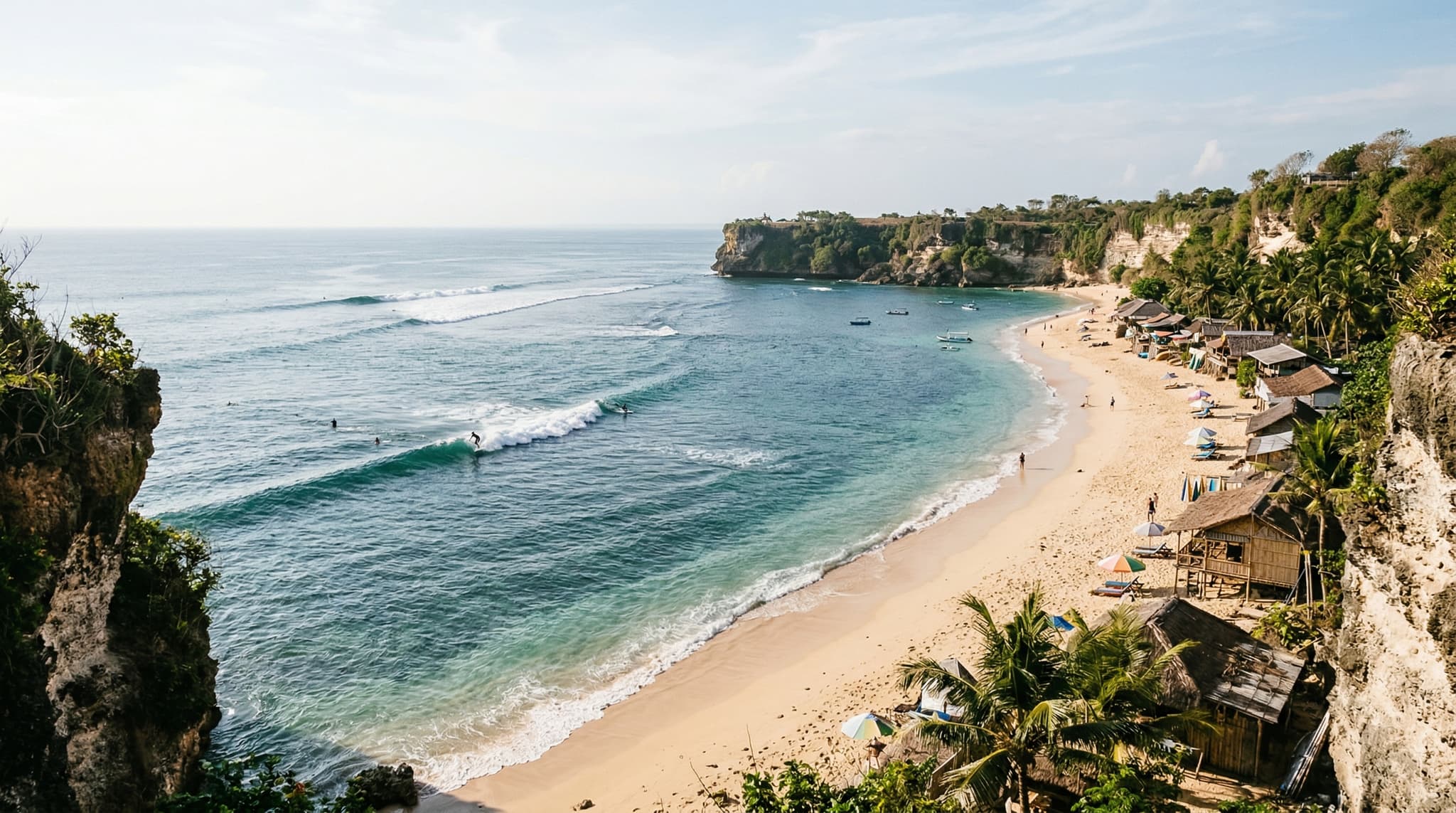 Balangan Beach on Bali's Bukit Peninsula — a crescent of golden sand framed by limestone cliffs, with turquoise water and a surfer riding the left-hand reef break in the foreground. Establishes the beach's dramatic setting and surf culture for this guide.