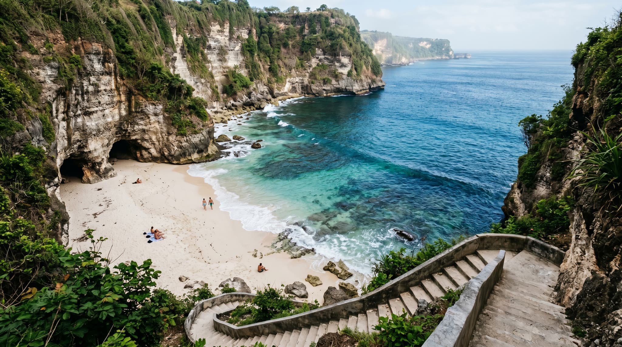 Green Bowl Beach on Bali's Bukit Peninsula — a narrow white-sand cove enclosed by towering limestone cliffs, with shallow turquoise water and the dark mouths of sea caves visible at the base of the rock face, conveying the remote, enclosed character of this cliff-access beach
