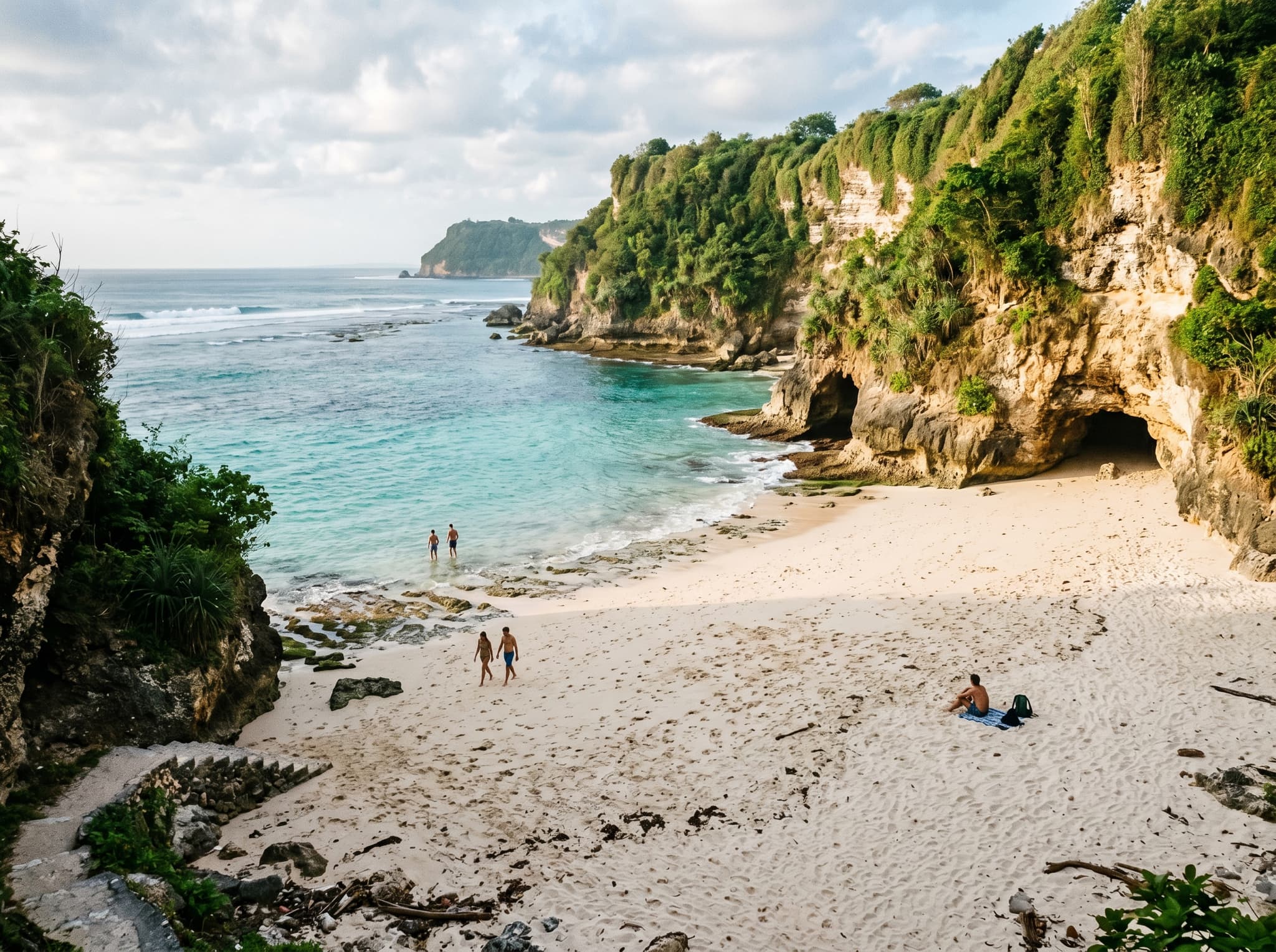 Green Bowl Beach at low tide from the sand level — the full width of the cove open, white sand stretching toward the water, limestone cliffs rising on both sides, a few visitors swimming or sitting near the cave entrances, showing the beach at its most accessible and rewarding