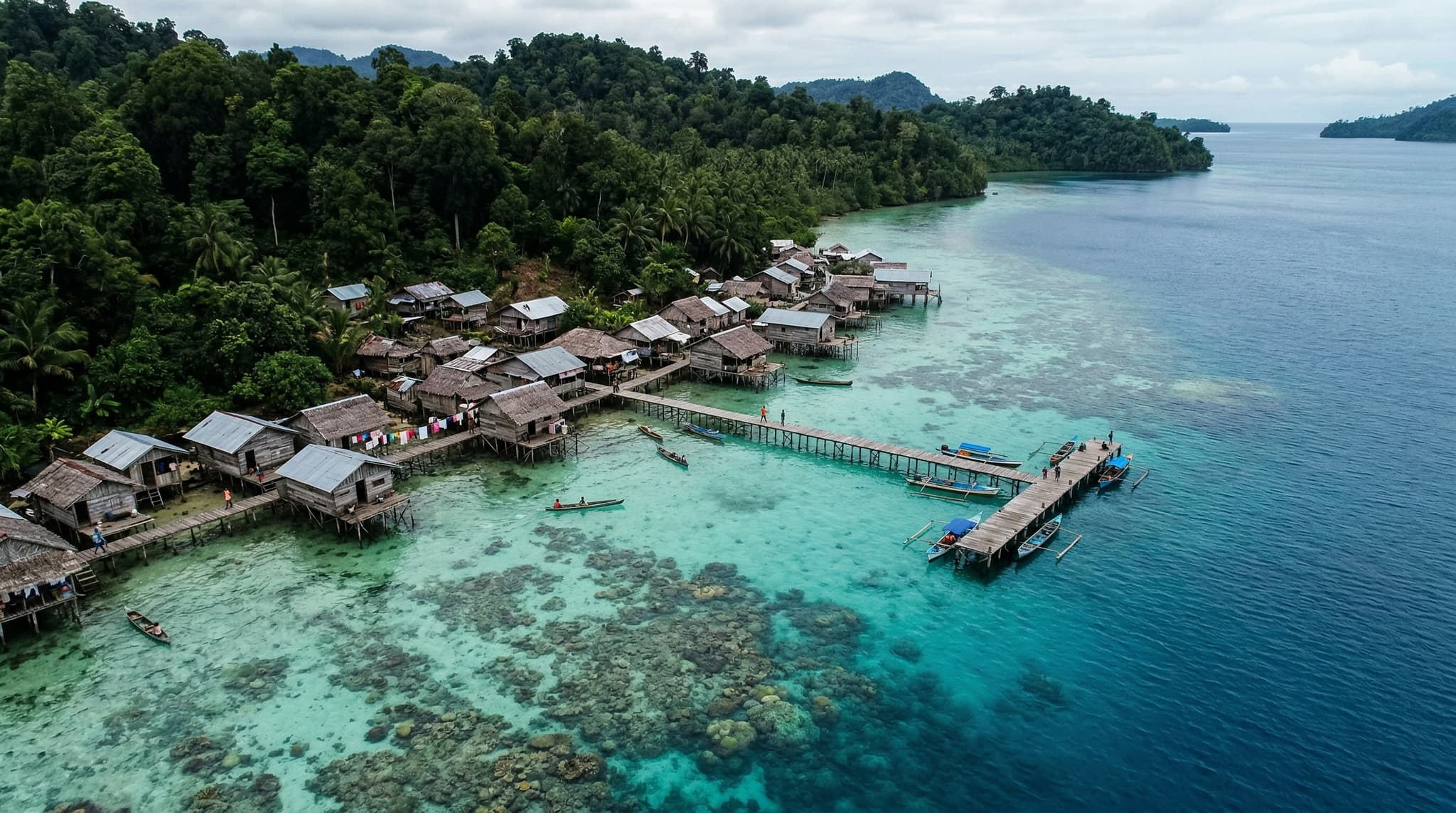 Aerial or water-level view of Sawandarek village on Mansuar Island, Raja Ampat — wooden stilt houses along the shoreline with dense tropical forest behind and vivid turquoise reef water in the foreground, establishing the article's central theme of a community living directly alongside its reef