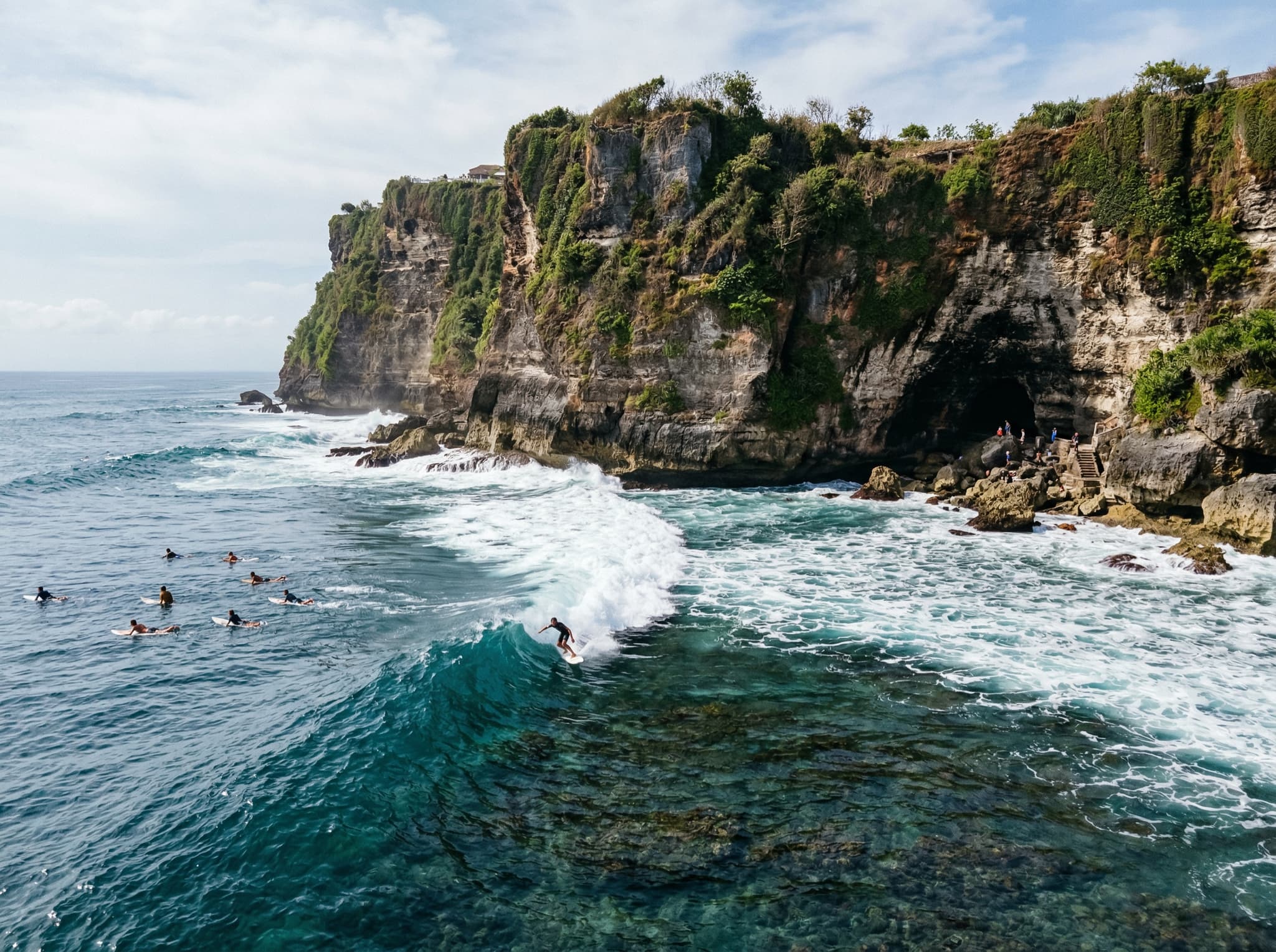 Uluwatu Beach surf break viewed from above or at sea level, showing the famous reef break with surfers in the water and the dramatic cliff backdrop — illustrating the nearby surf destination mentioned in the 'What's Nearby' section