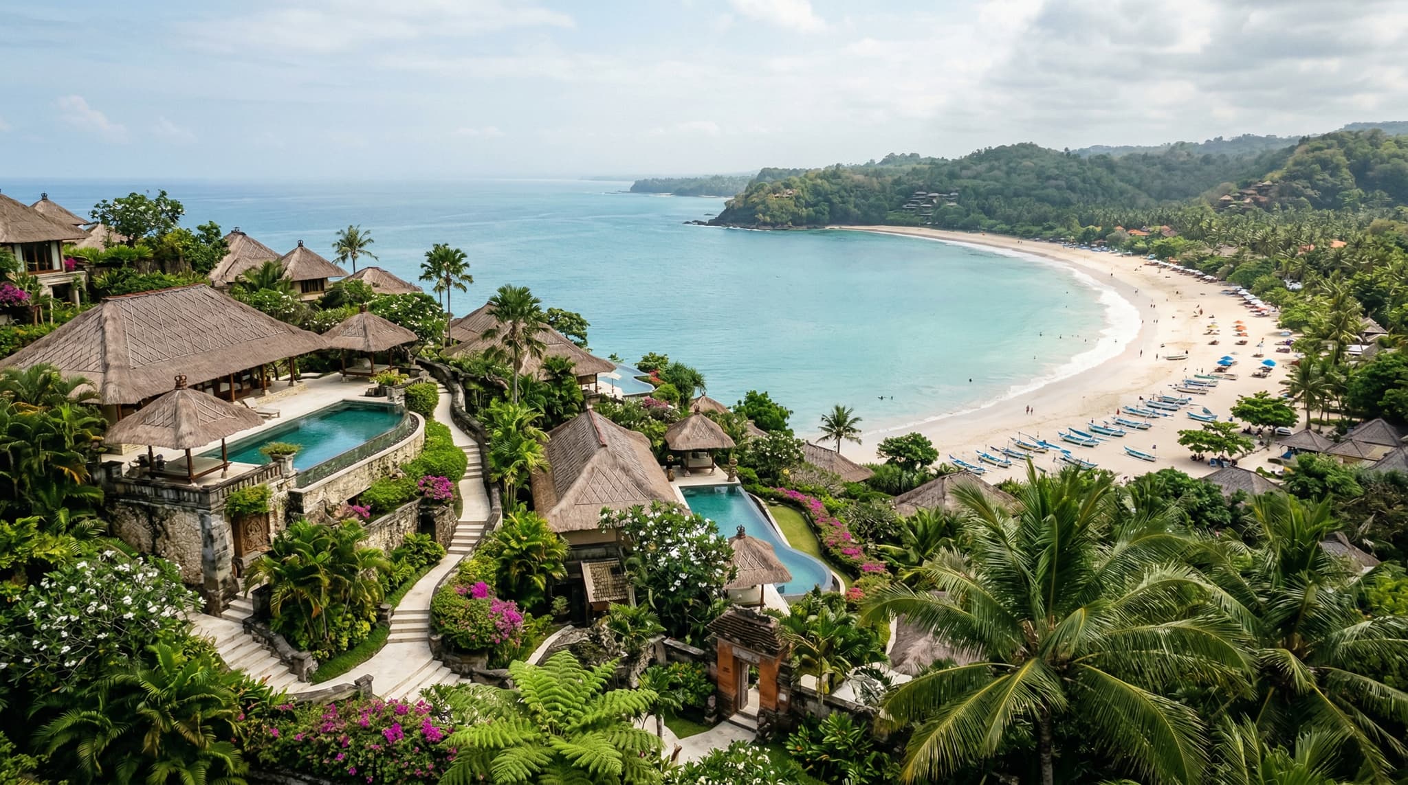Aerial or elevated view of Four Seasons Jimbaran Bay resort terraced into the hillside above Jimbaran Bay, with traditional thatched-roof Balinese villa compounds visible among tropical gardens, the curved bay and turquoise water in the background — establishing the resort's quiet, residential character described throughout the article