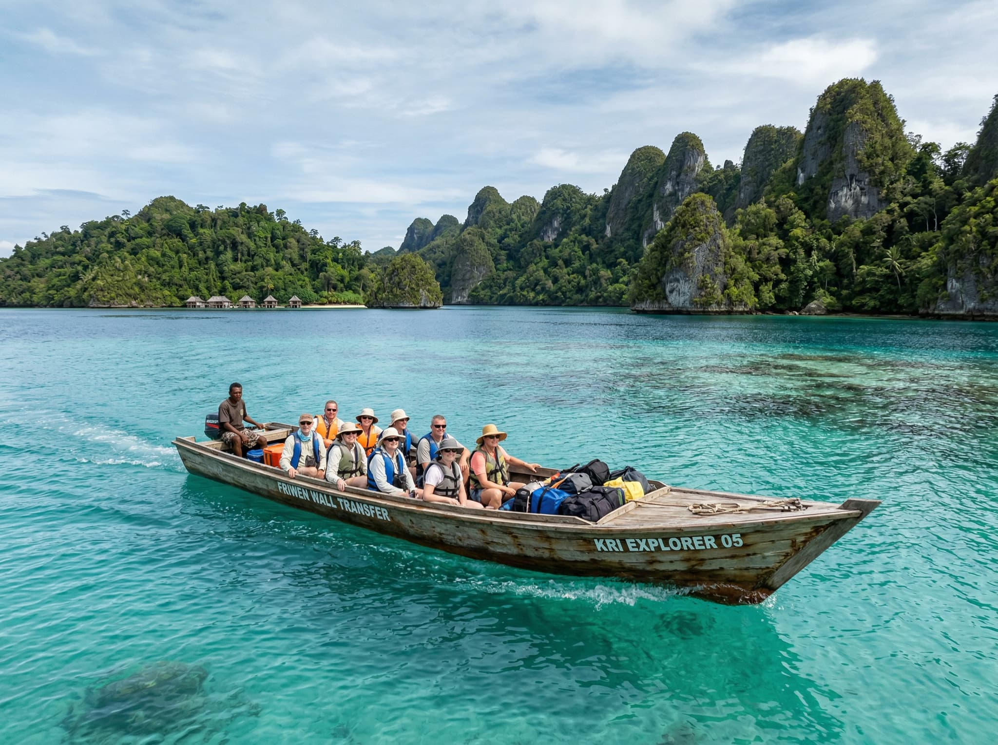 A wooden speedboat crossing calm turquoise water between islands in Raja Ampat, with forested limestone karst islands visible in the background — illustrating the boat transfer journey from Kri Island or Waisai to reach Friwen Wall.