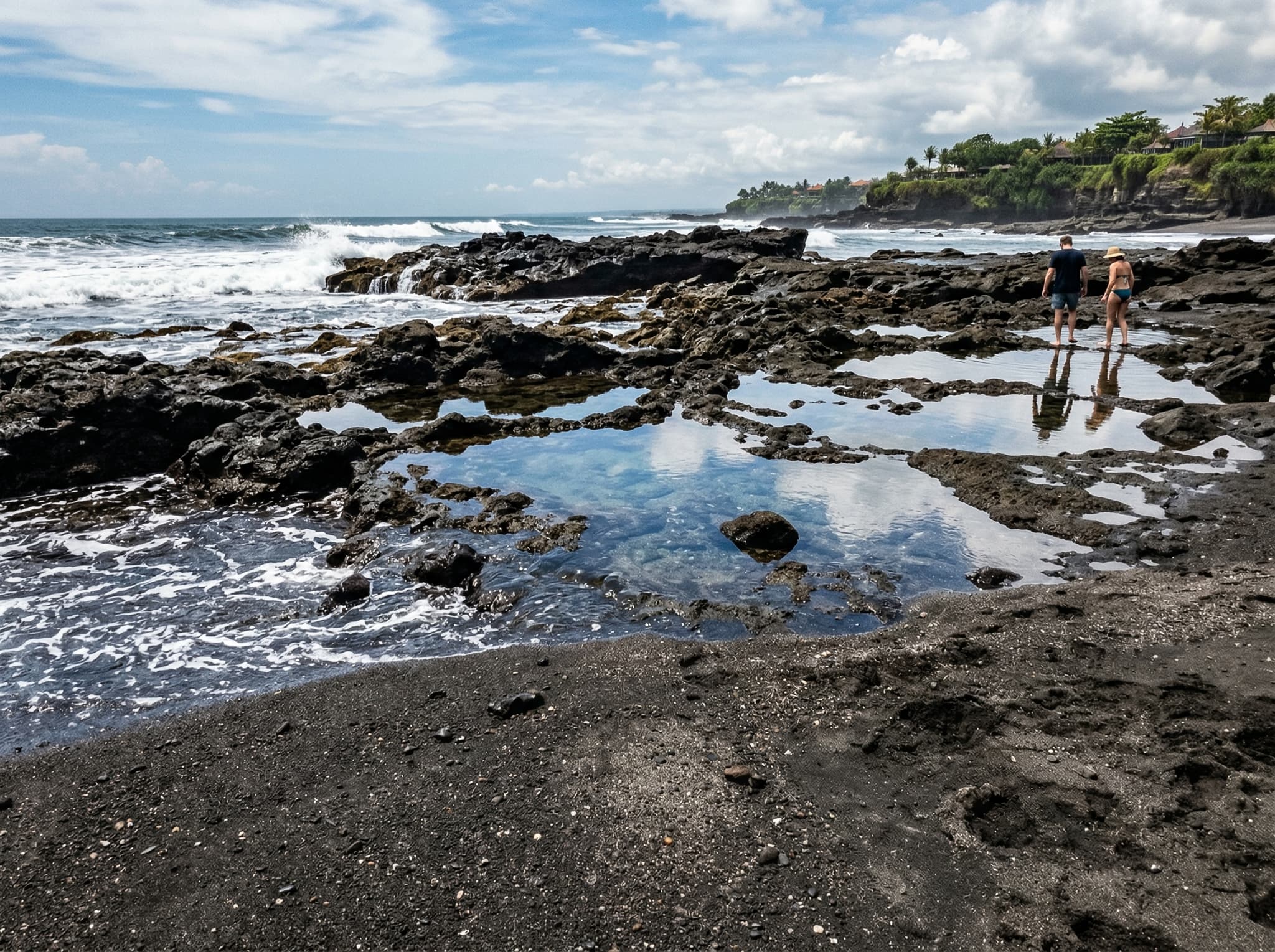 Close-up of Echo Beach's distinctive black volcanic sand and rocky shoreline at low tide, showing the coarse dark texture and rock pools that characterize this end of Canggu's coastline — contrasting with the white-sand beaches of southern Bali