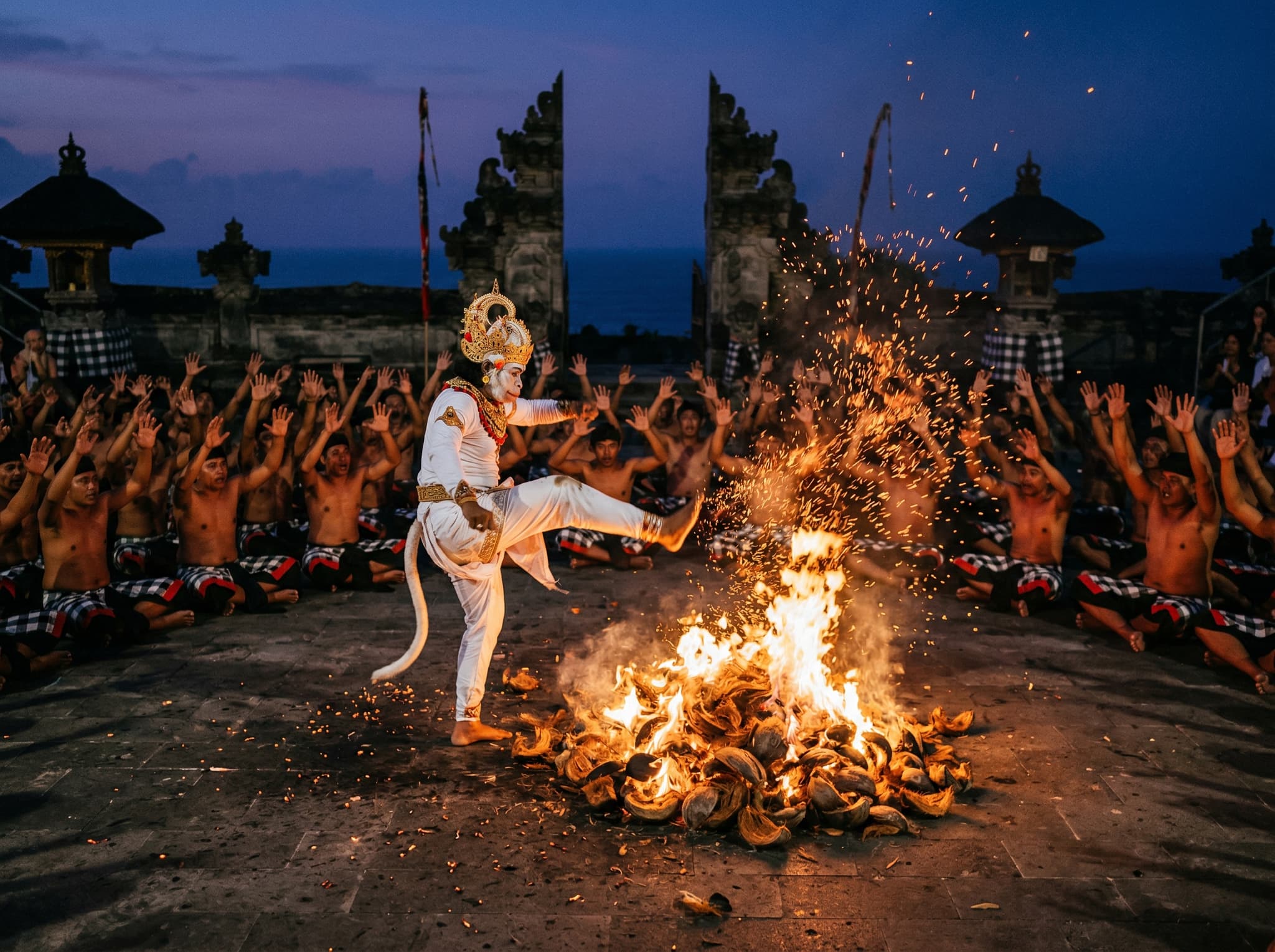 The Kecak fire finale — a performer in trance kicking through a pile of burning coconut husks, scattering embers across the stone stage at Uluwatu Temple amphitheater, representing the Ramayana episode in which Hanuman escapes execution by fire