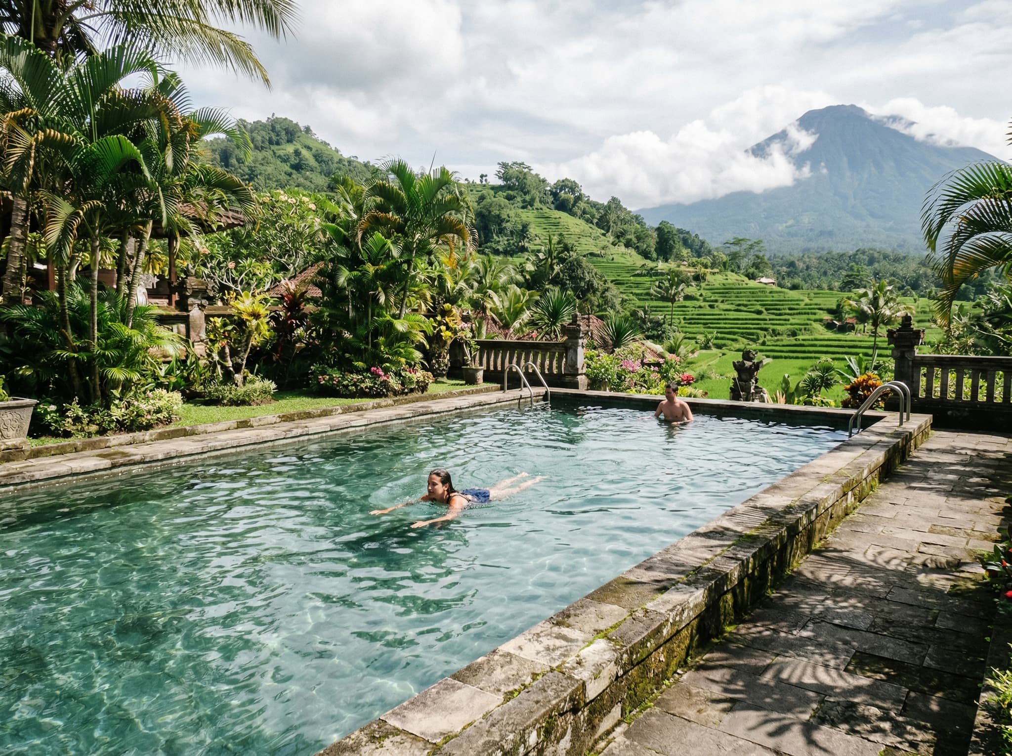 The spring-fed upper swimming pool at Tirta Gangga with views toward the surrounding gardens and the slopes of Mount Agung — illustrating the article's mention of the unexpected bonus of swimming in cool spring water with a volcanic backdrop.