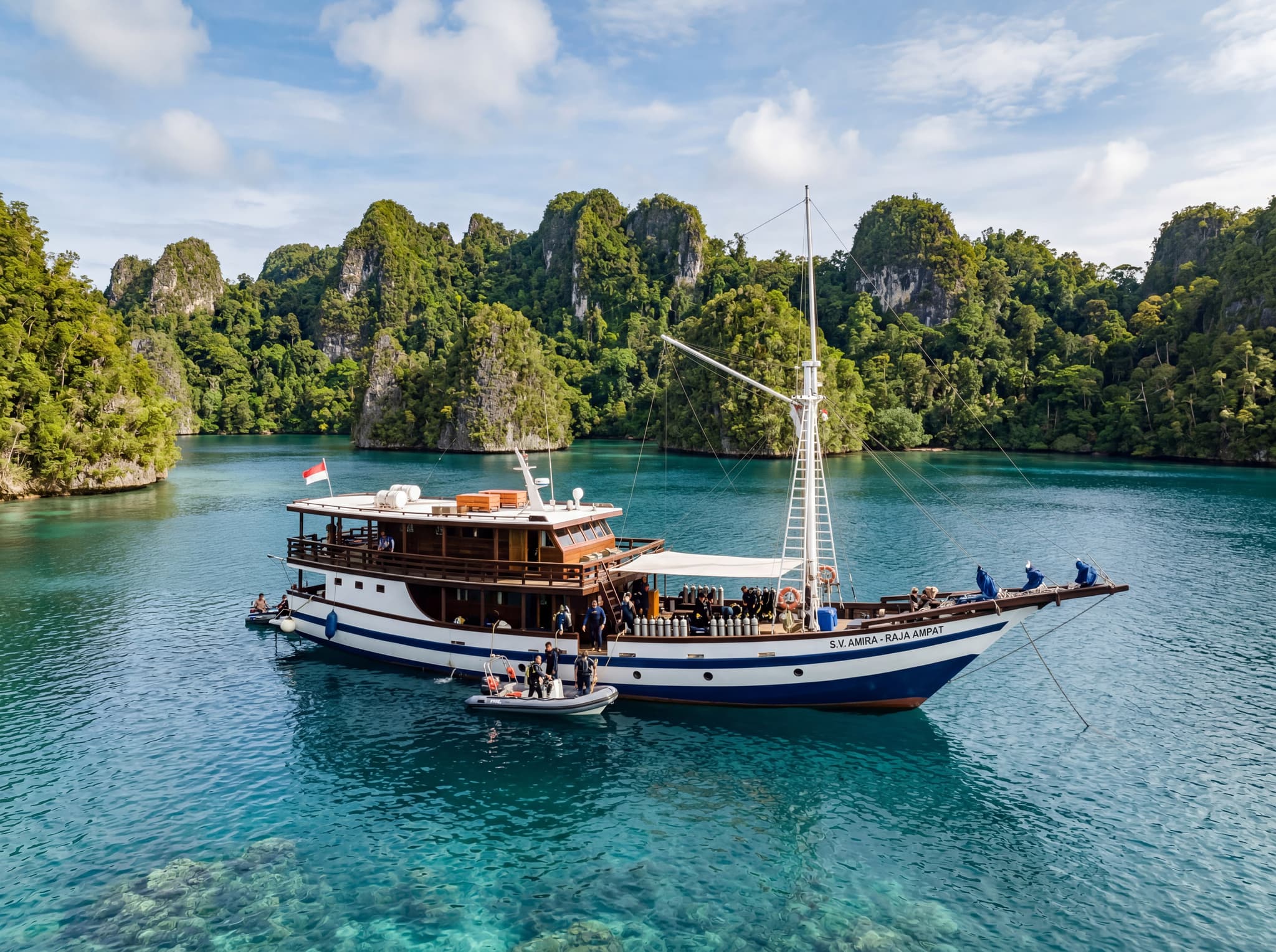 A liveaboard dive vessel anchored in calm blue water near a Raja Ampat island, with jungle-covered karst formations in the background — representing the primary access method to Sardine Reef described in the logistics section