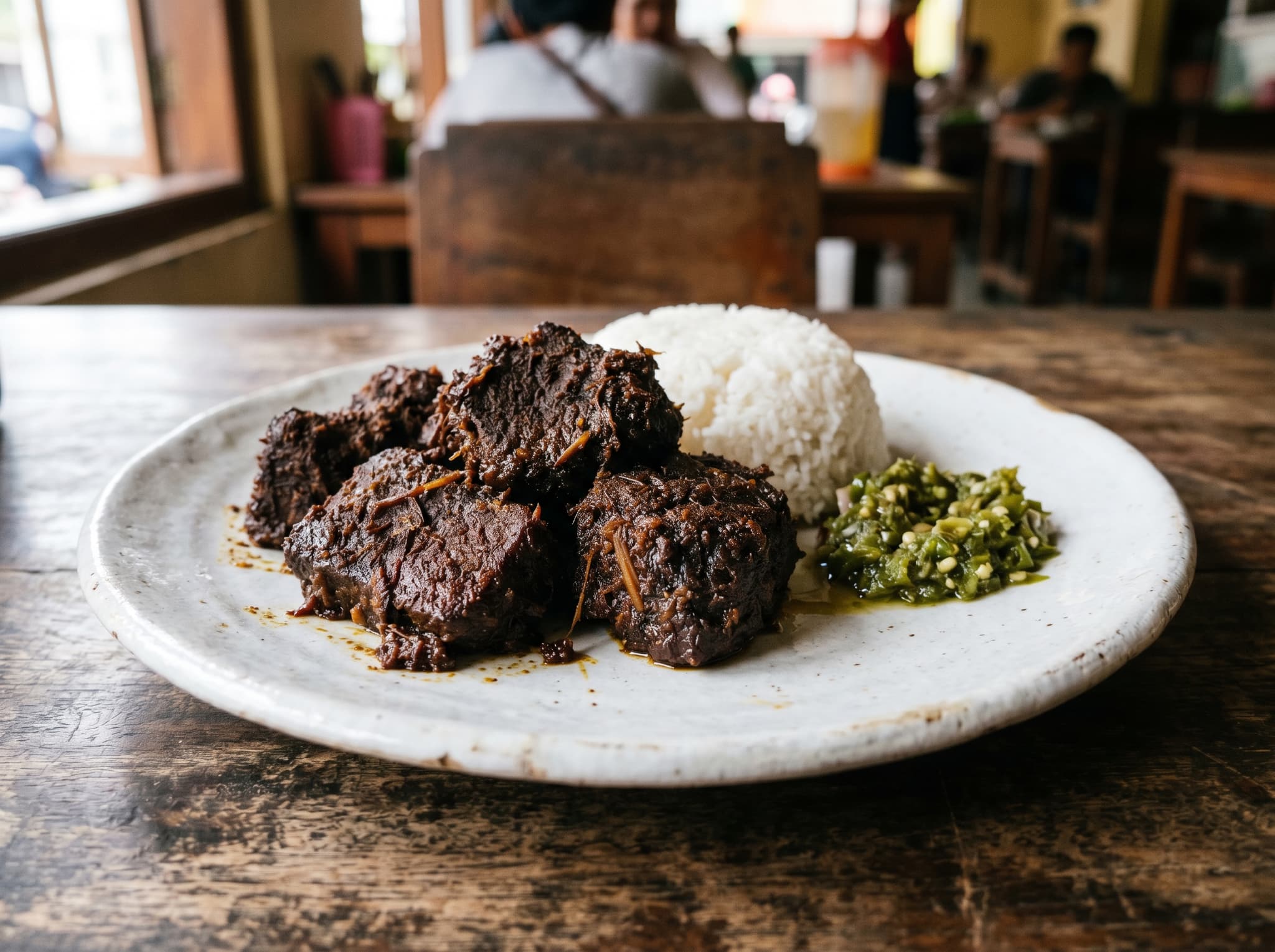 A close-up of rendang — dark, dry, intensely caramelized beef — on a white plate with a small mound of white rice and a green chili sambal, showing the concentrated texture that distinguishes it from a curry or stew. Central image for the Padang food section's most famous dish.