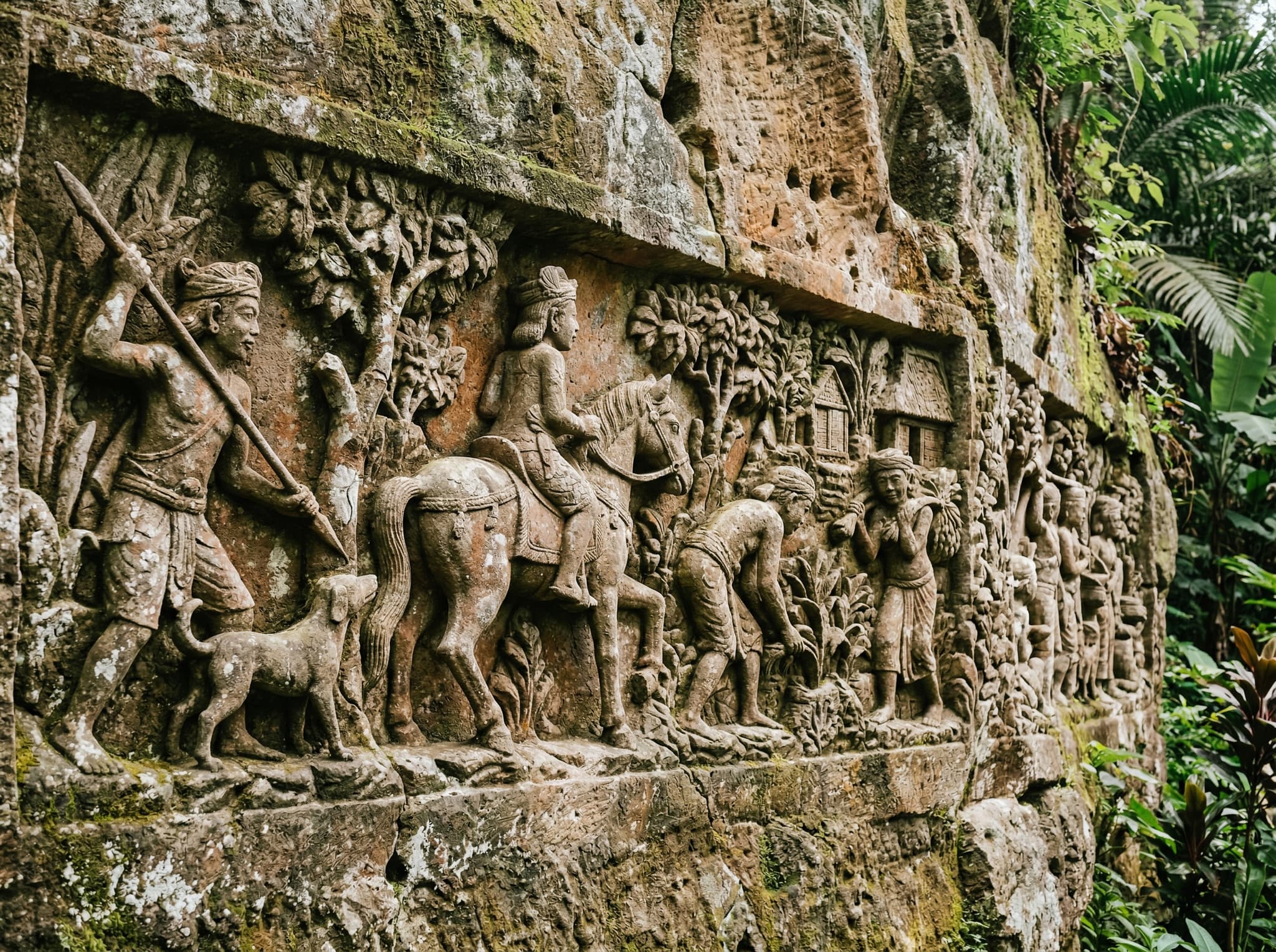 Close-up detail of carved figures on the Yeh Pulu relief — weathered stone panels showing scenes of village life, farming, and religious motifs from the 14th-century Kingdom of Pejeng, illustrating the narrative quality of the carvings described in the article