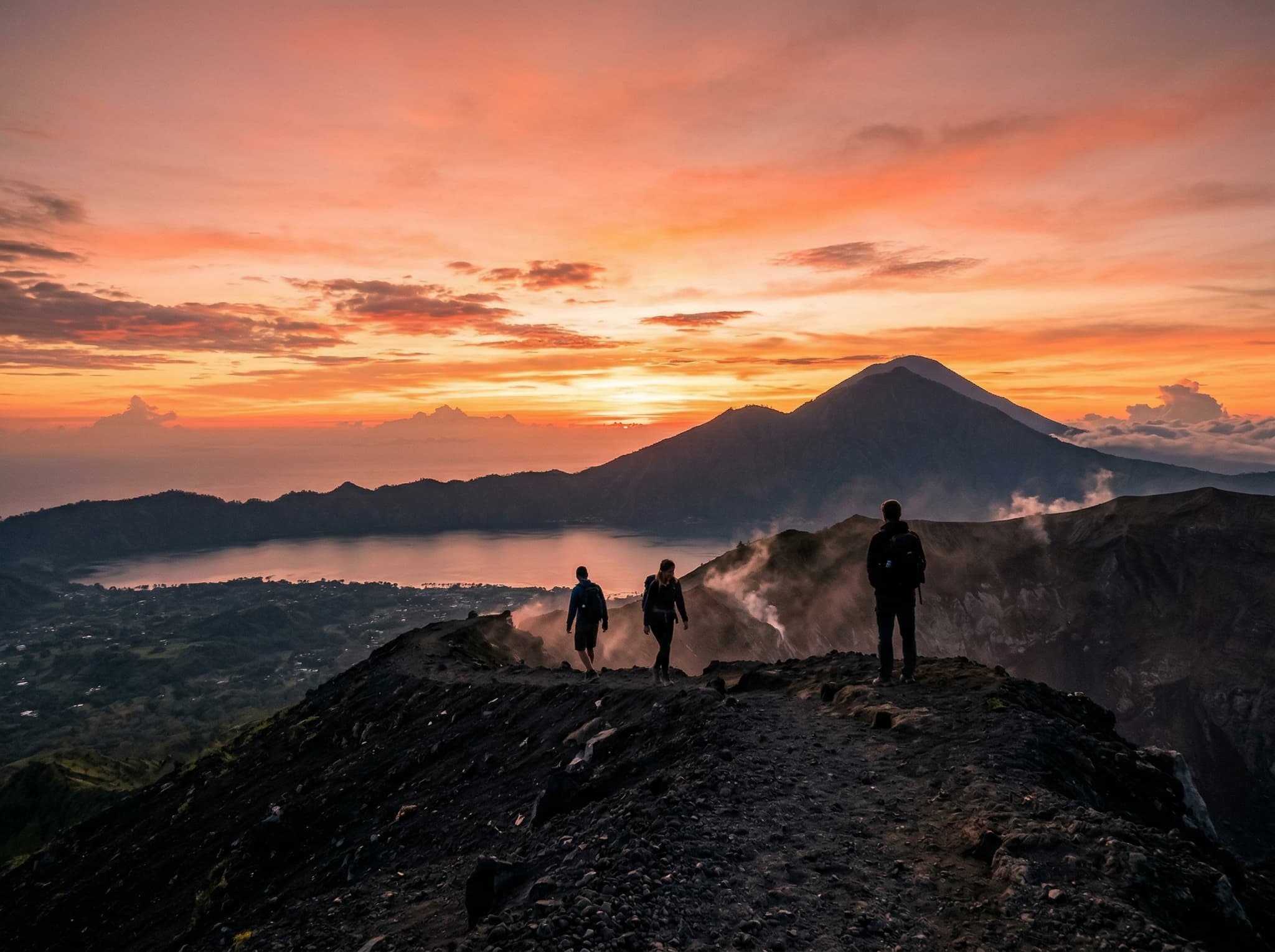 Mount Batur summit at sunrise with trekkers silhouetted against the orange and pink sky, Mount Agung visible in the distance — capturing the iconic sunrise trek experience that is the headline activity of a Kintamani visit