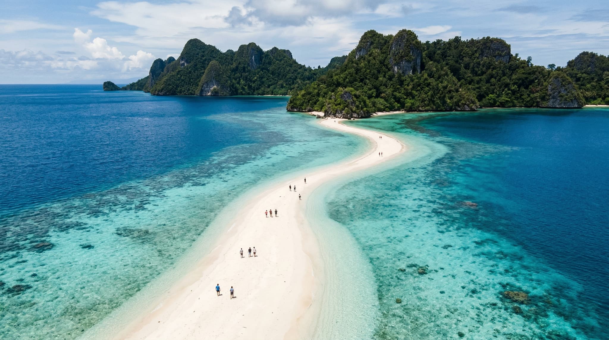 Pasir Timbul sandbar emerging from turquoise water in Raja Ampat, Indonesia — a narrow white sand ridge surrounded by clear sea with karst limestone islands rising on the horizon, showing the tidal phenomenon at peak low tide