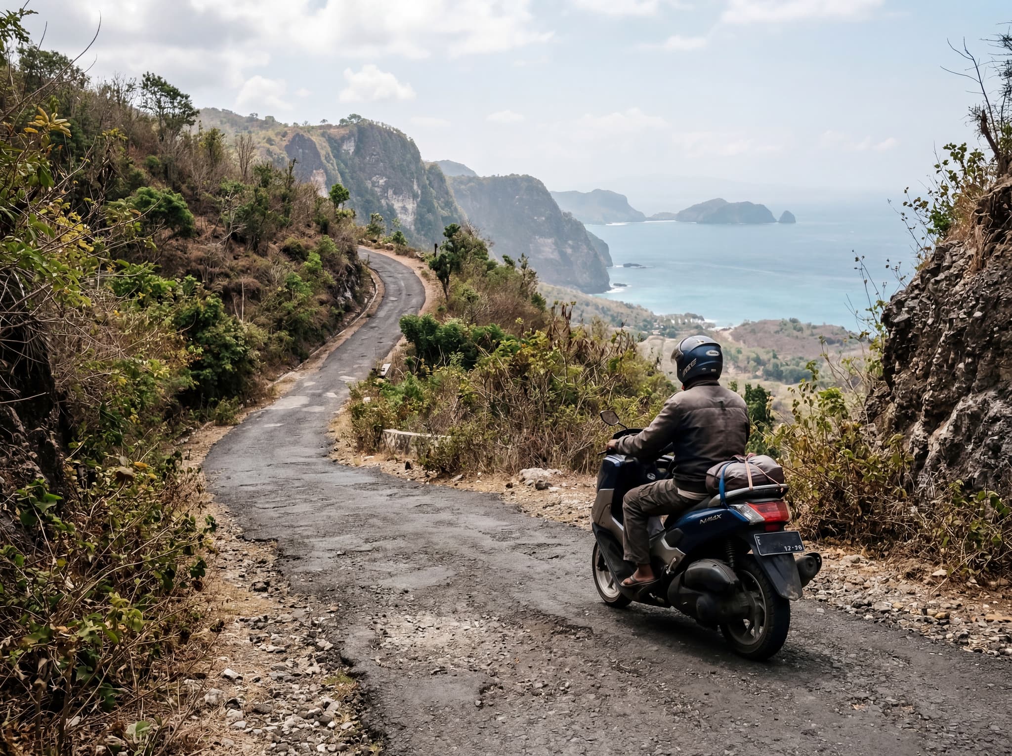 A local driver or scooter rider navigating a steep, narrow road on Nusa Penida with lush hillside or cliff scenery visible — illustrating the article's warning about the island's challenging road conditions and the recommendation to hire local drivers