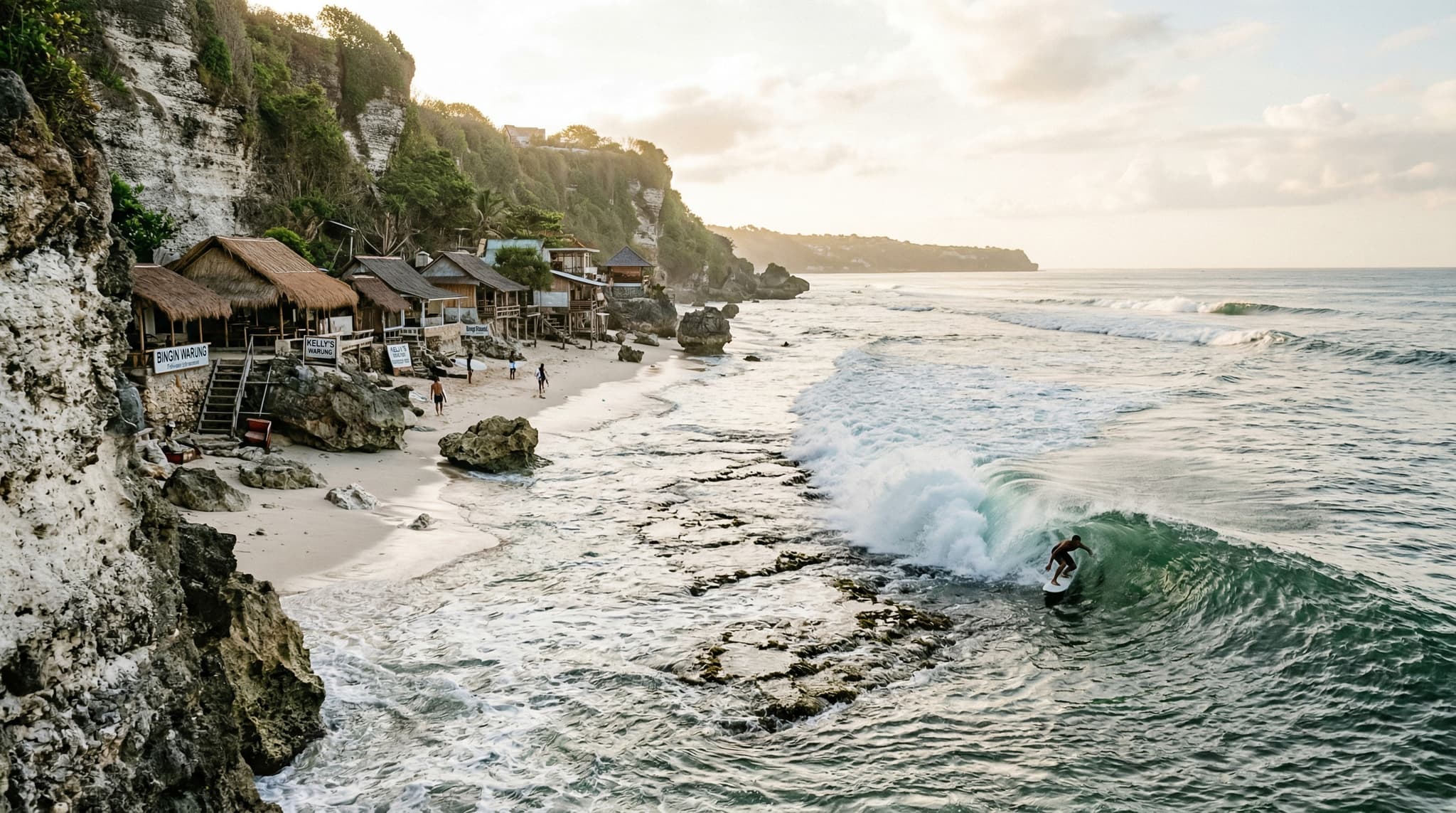 Bingin Beach on Bali's Bukit Peninsula viewed from the base of the limestone cliffs, showing the narrow white sand beach, turquoise water, and a surfer riding a left-hand barrel over the shallow reef — establishing the beach's dramatic setting and surf culture identity