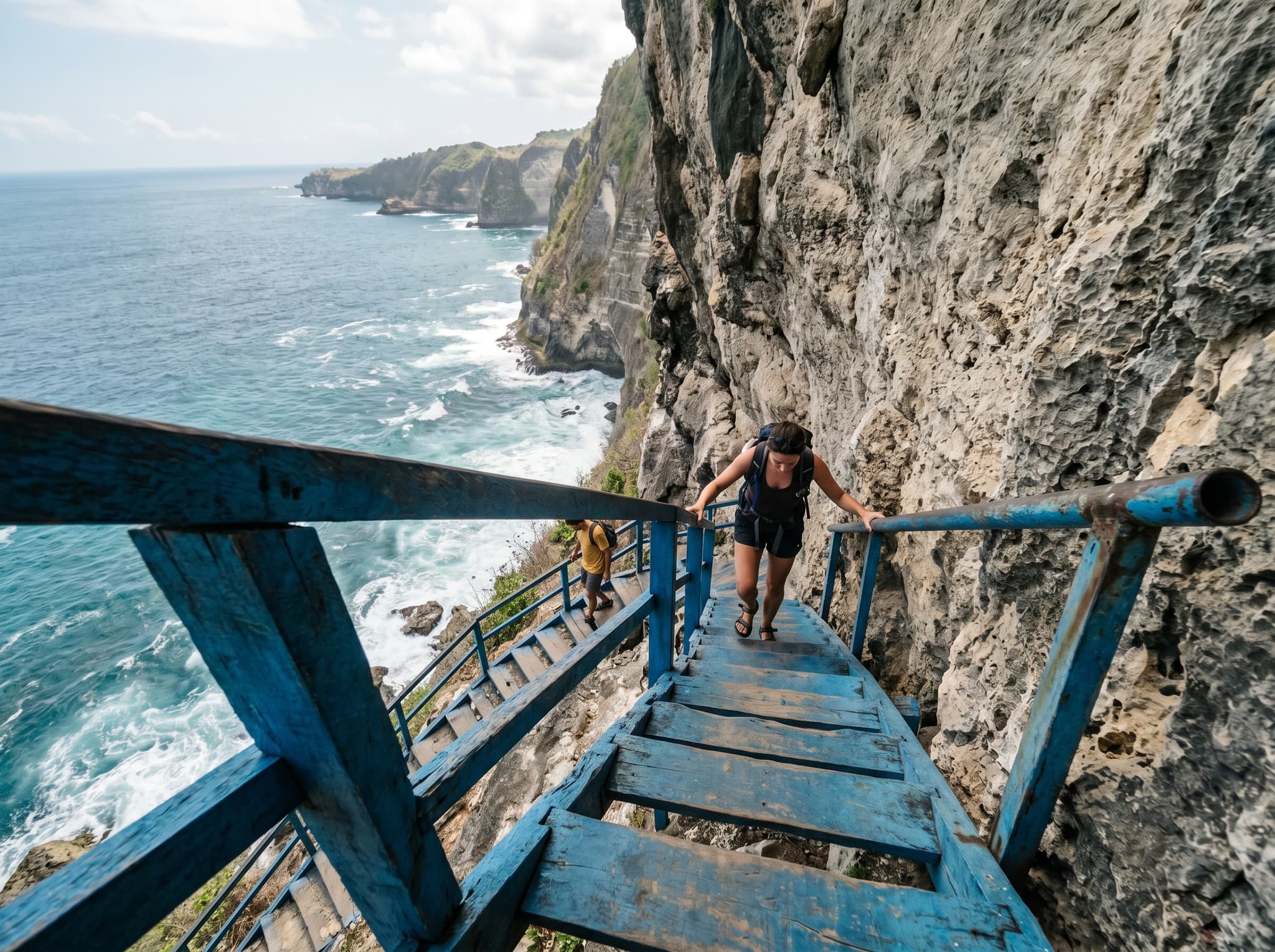 A close perspective on the Peguyangan blue staircase mid-descent — narrow wooden steps fixed to the cliff face, some sections without railings, the sheer drop visible to one side and the ocean below. Captures the physical reality of the descent described in the article: steep, exposed, and demanding.