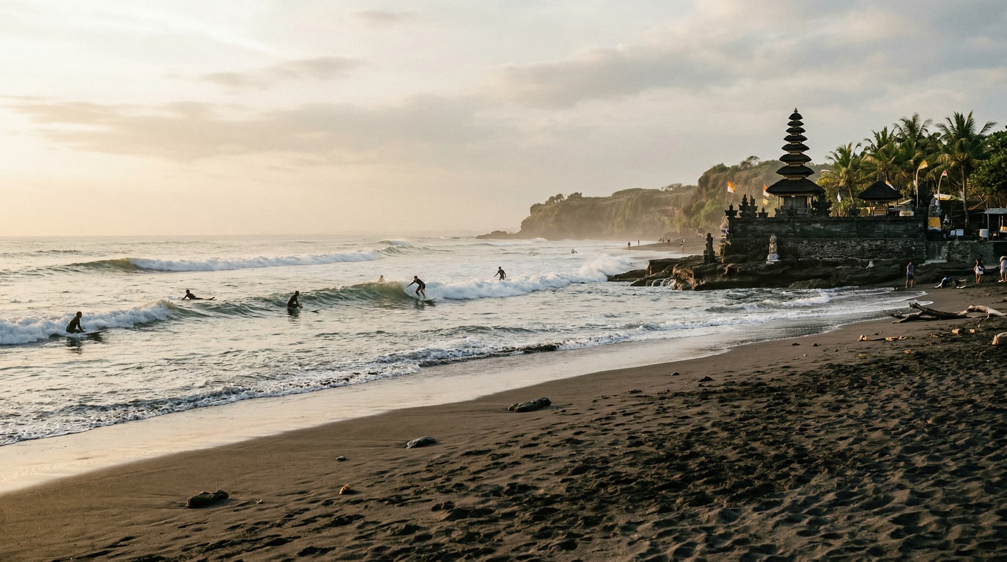 Pererenan Beach, Bali — dark volcanic black sand shoreline with consistent surf waves breaking in the middle distance, Pura Masceti temple silhouette visible at the water's edge, warm late-afternoon light, a handful of surfers in the lineup, low coastal cliffs in the background. Establishes the beach's moody, uncrowded character central to the article's premise.