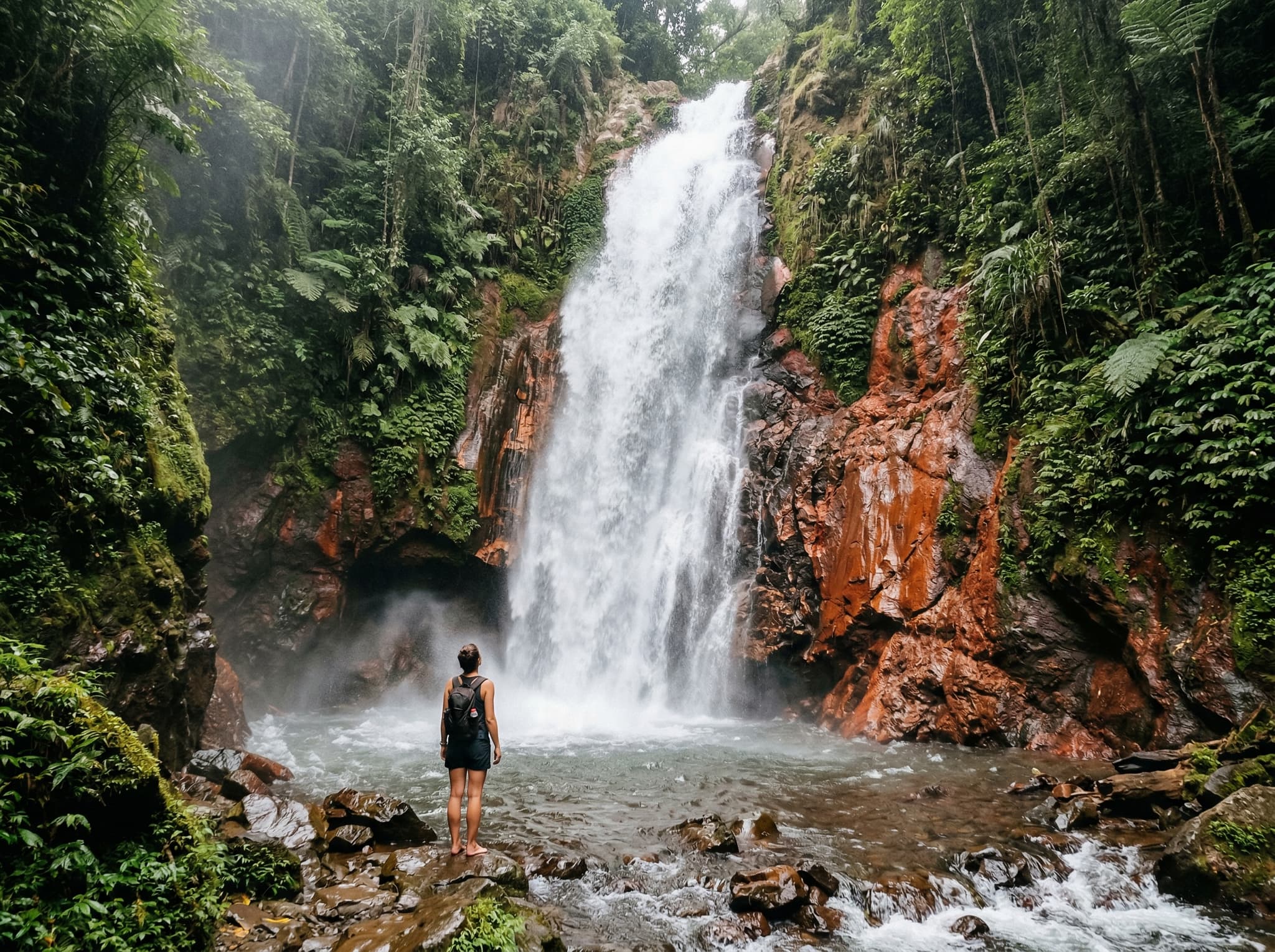 A lone traveler standing at the base of Labuhan Kebo waterfall in the shallow pool, surrounded by jungle with no other visitors in sight — capturing the rare solitude that makes this waterfall worth the effort over Munduk's more crowded alternatives