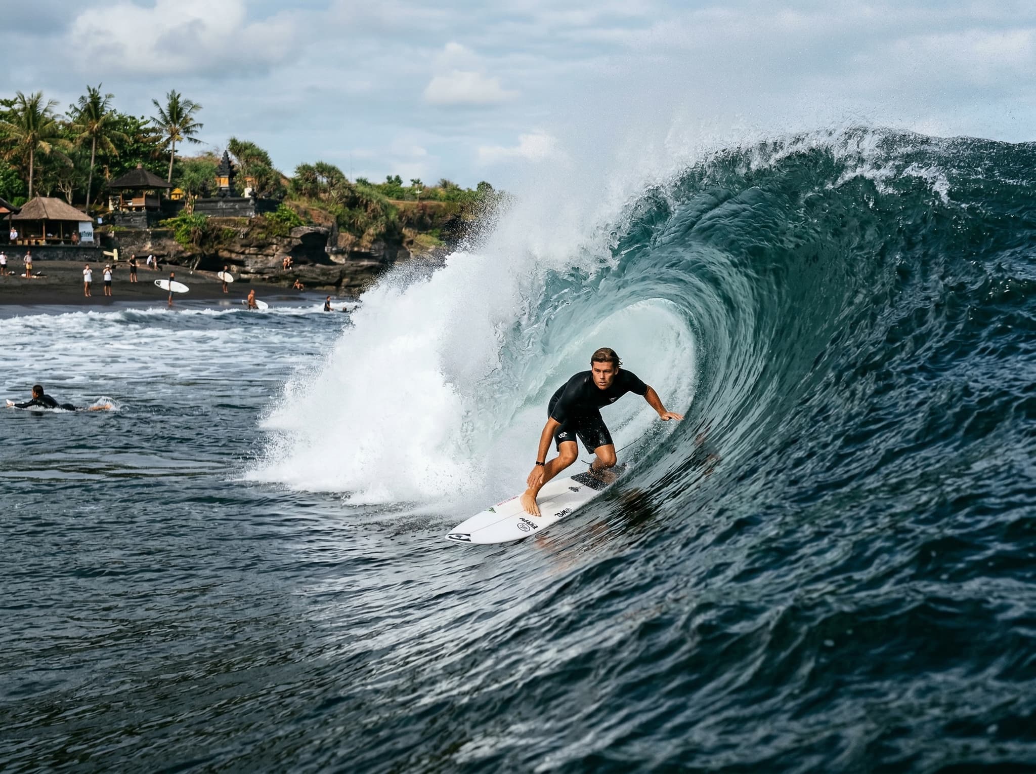A surfer riding or paddling out at Keramas Beach's powerful right-hand reef break, with the dark water and volcanic sand visible — illustrating the wave quality that earned Keramas its WSL Championship Tour reputation