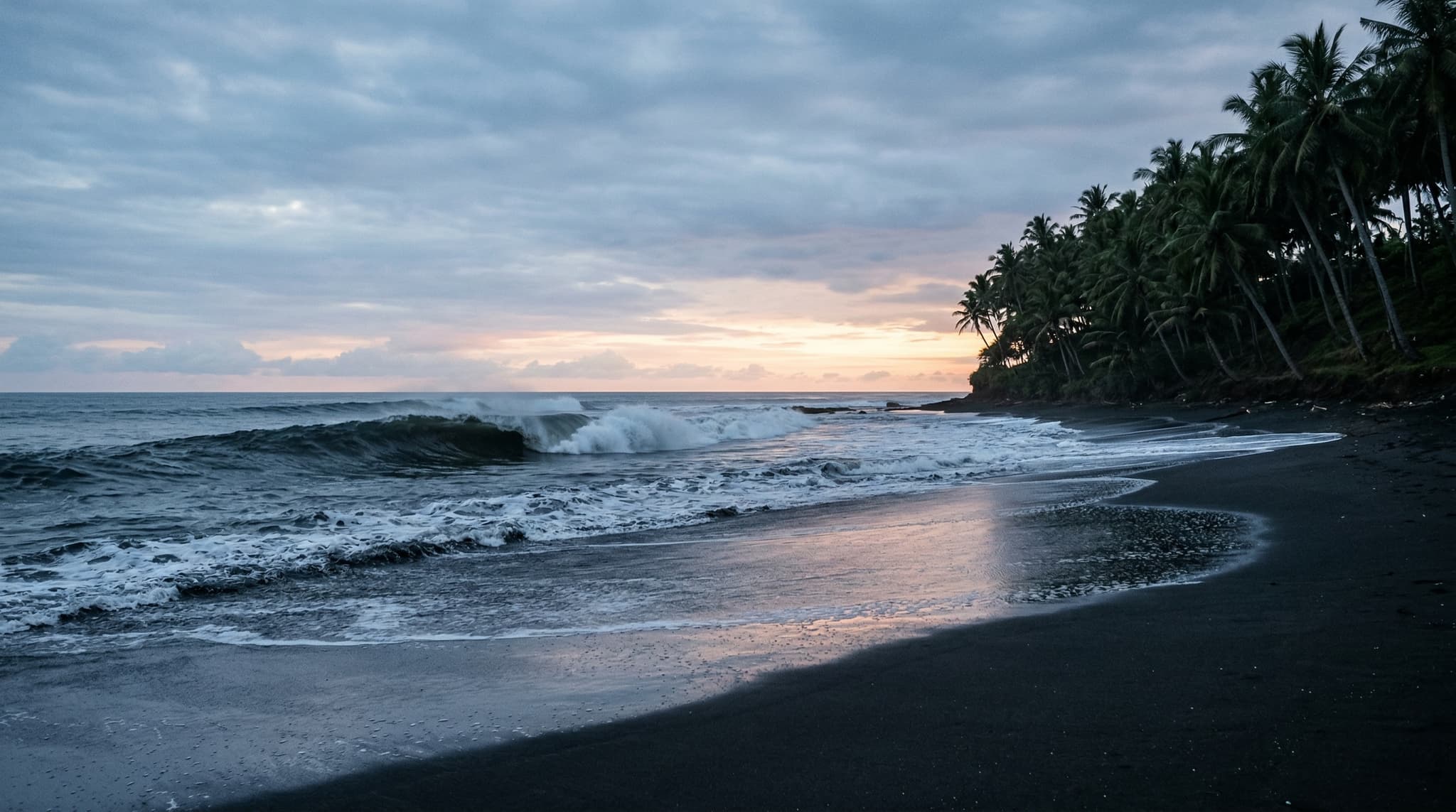 Keramas Beach's distinctive black volcanic sand shoreline at dawn or early morning, with dark surf breaking over the reef and coconut palms visible along the coast — establishing the raw, unpolished character of this East Bali surf destination